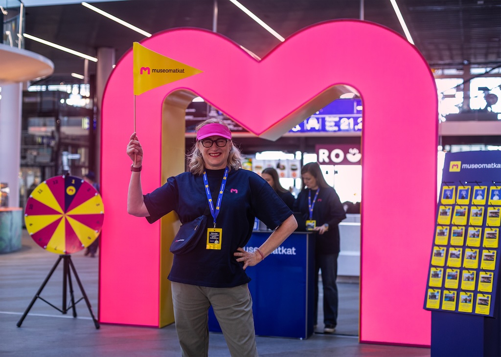 Smiling woman in a pink cap and navy shirt poses with a yellow flag under a large pink heart arch at an event booth.