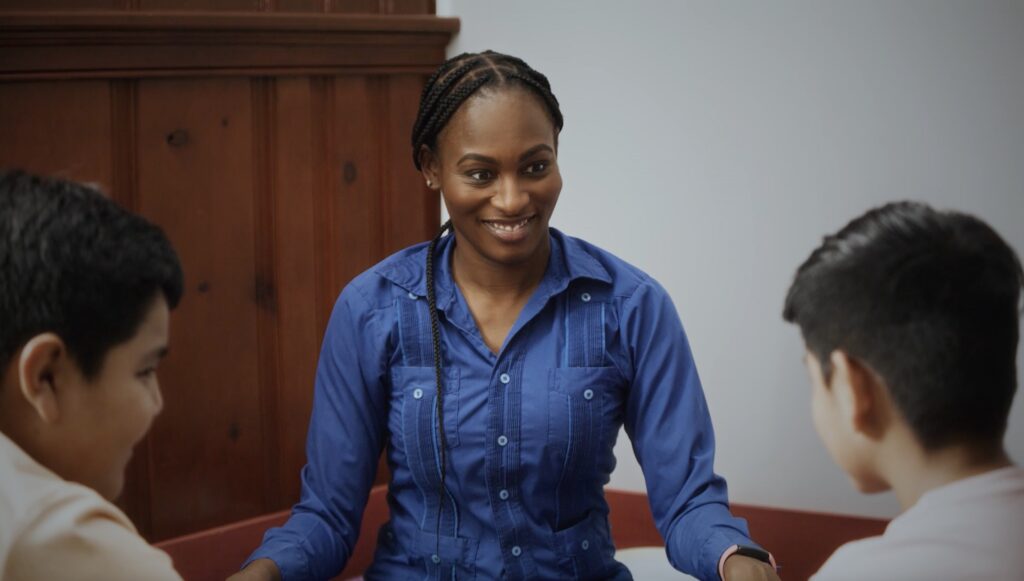 Screenshot | Muse by Clios A smiling woman with braided hair and a blue button-down shirt leads a discussion at a table with two seated coworkers in a meeting or classroom setting.