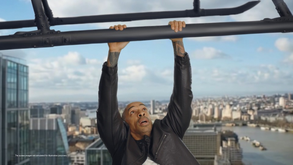 Man in a leather jacket hangs from a high horizontal bar above a city skyline with tall buildings in the distance.