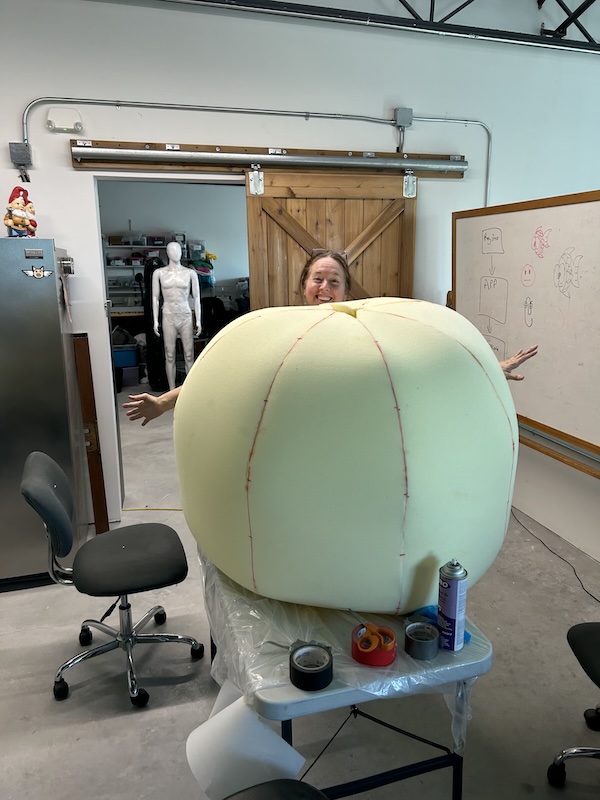 Person smiling behind a giant pale-yellow peach-shaped prop on a wheeled table in a workshop.