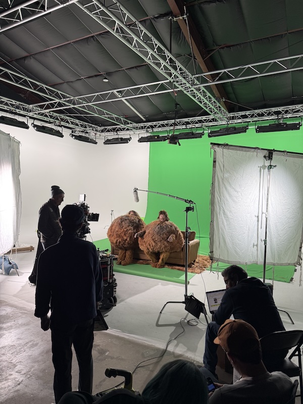 Film set with a green screen wall, crew filming two performers in large furry costumes on a couch.