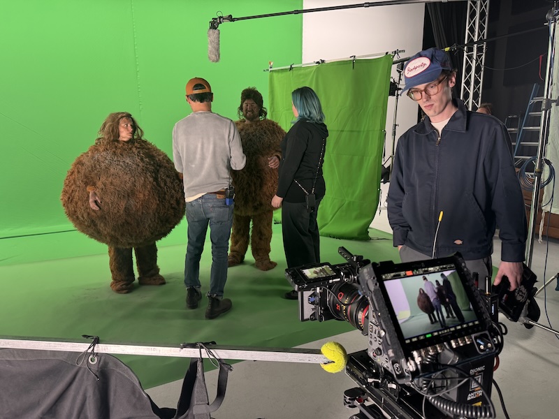 Behind-the-scenes on a green-screen set: crew members film two people in large furry monster costumes with a camera and boom mic nearby.