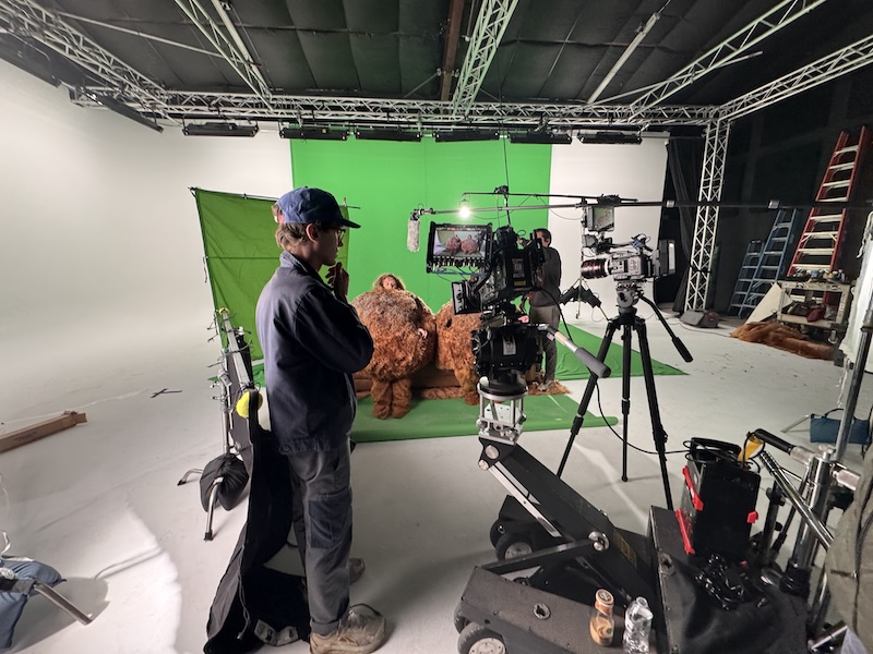 Film studio with a bright green chroma key backdrop, camera rig on a dolly, and crew members adjusting gear while two large plush teddy bears sit in front of the screen behind them.