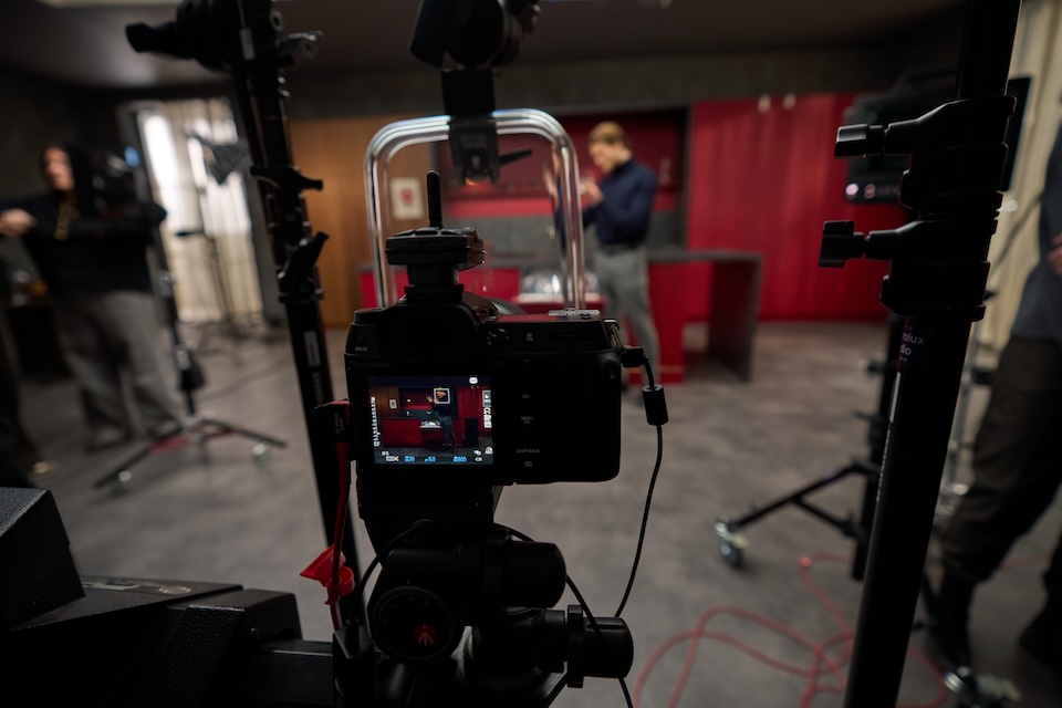 Professional camera on a tripod in a studio, facing a red-curtained set with crew in the background, blurred.