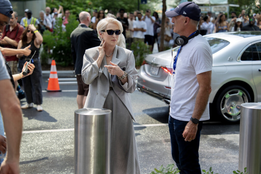 Woman in a light gray suit and sunglasses talks to a man wearing a cap and headphones near a silver car, with a crowd in the background.