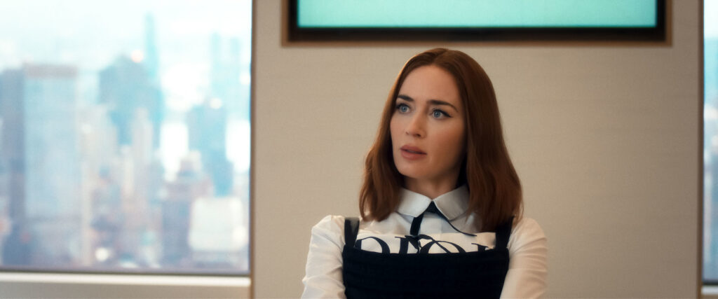 Woman with auburn shoulder-length hair sits in a bright office near a window, wearing a white blouse and black dress, looking to the side with a city skyline blurred in the background.