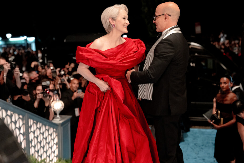 Woman in a bright red gown smiles at a man in a suit on a blue carpet as photographers crowd behind them at night festival of red carpet events.