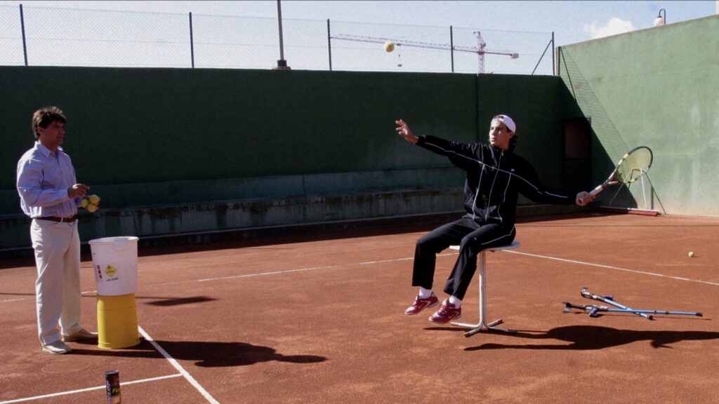 nadalwithuncle | Muse by Clios Tennis practice on a clay court: a player in a black tracksuit serves a ball while a coach stands nearby with a bucket of balls and tennis gear.",