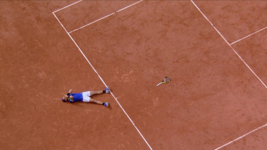 nadaloncourt | Muse by Clios Overhead view of a clay tennis court with a player in blue shirt and white shorts lying on the court beside a fallen racket.