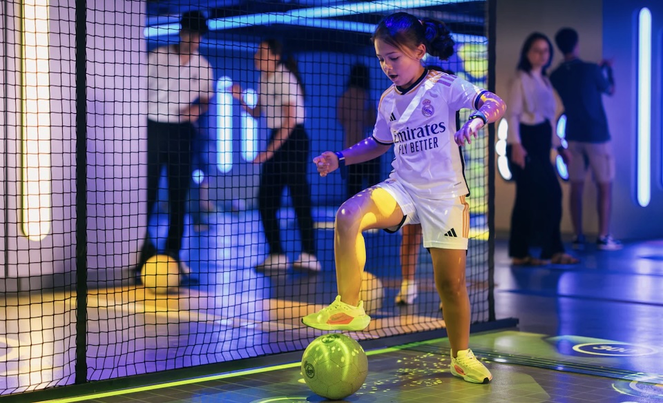 Young girl in a Real Madrid jersey balances on one foot, kicking a neon-green ball inside an indoor futsal arena with netted walls and spectators in the background