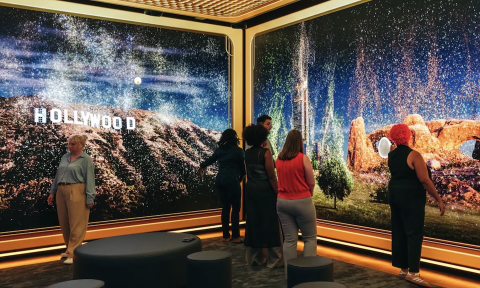Visitors stand in a bright exhibition room as a wide wall mural shows a Hollywood landscape with the sign and a starry sky across curved screens.
