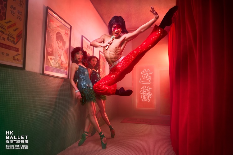 Male dancer leaps high in red glitter pants backstage, microphone in hand, as three women in sequined dresses watch.