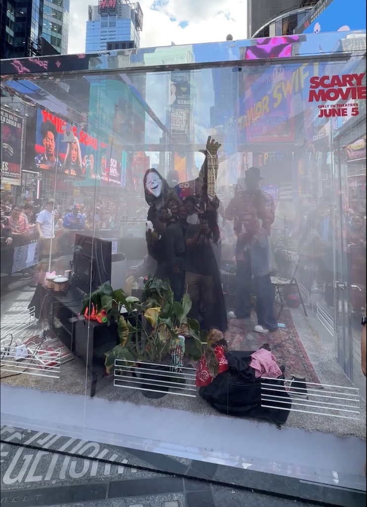 Screenshot | Muse by Clios Glass storefront display in Times Square reflecting passersby; a masked performer sits inside among plants and props.