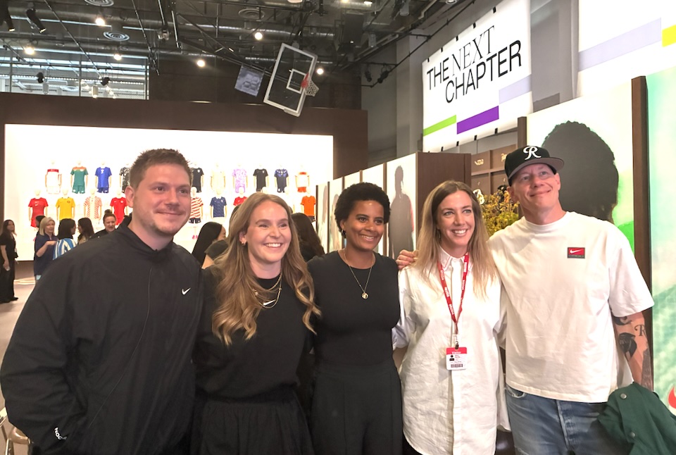 Group of five smiling attendees posing at a fashion expo booth, with a prominent'THE NEXT CHAPTER' sign in the background.