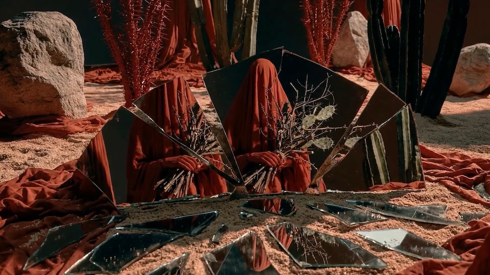 Art installation with a red-cloaked figure surrounded by broken mirrors on sandy ground, desert plants and rocks.