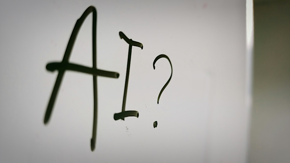 Close-up of dark green graffiti on a pale surface reading 'AI?' with a large A and I, followed by a question mark on glass or window