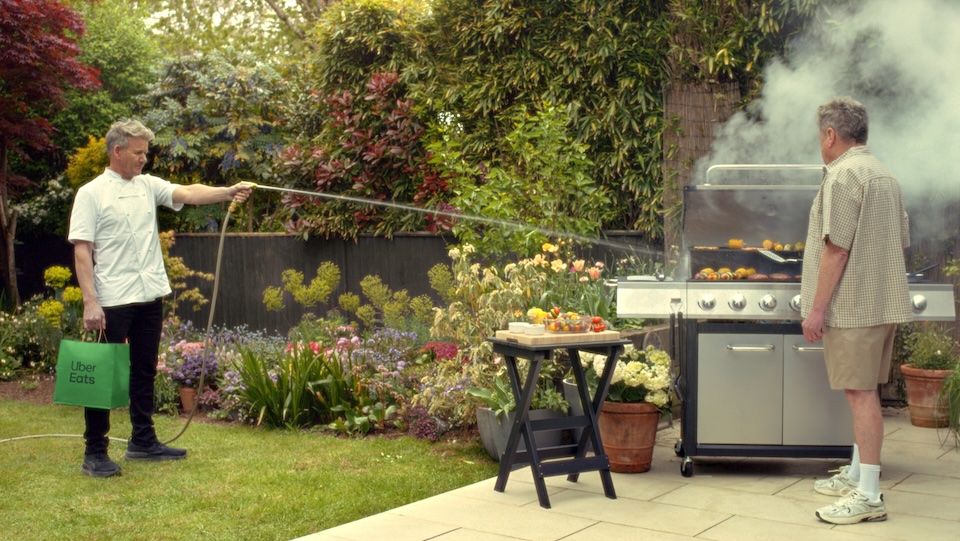 3616x2 - 1 | Muse by Clios Man in a white shirt holds a hose toward a large gas grill while another man stands at the grill in a garden backyard; Uber Eats bag in the foreground.