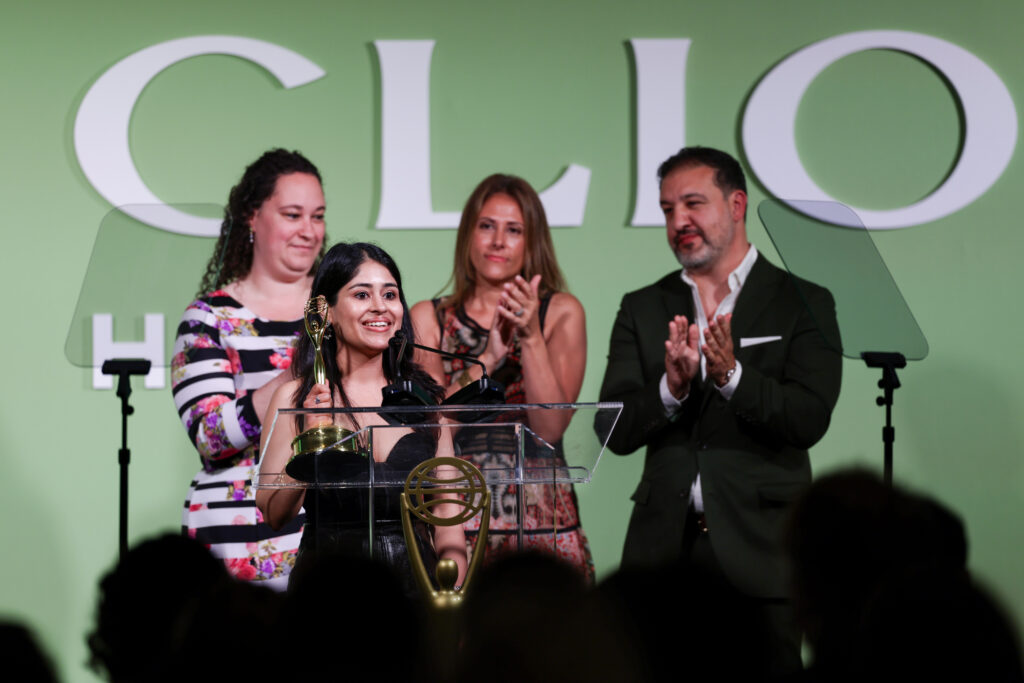 Woman at a clear podium accepting a trophy while smiling, with three people applauding behind her on a green stage backdrop.