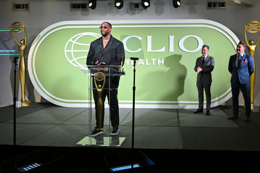Speaker at a clear podium on a curved green stage backdrop that reads ECLO Health, with two suited men applauding to the right and gold statues on either side of the scene.