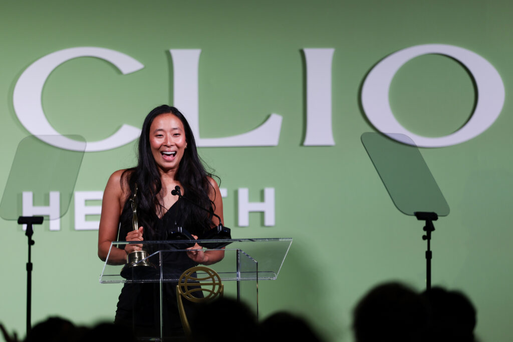 Woman on stage at an awards ceremony, smiling while holding a trophy behind a clear podium with a green backdrop.