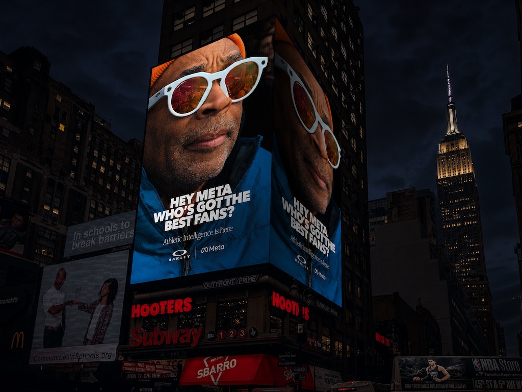 Nighttime city scene with a giant billboard of a man in white sunglasses and an orange beanie, captioned: 'Hey Meta, who’s got the best fans?'.