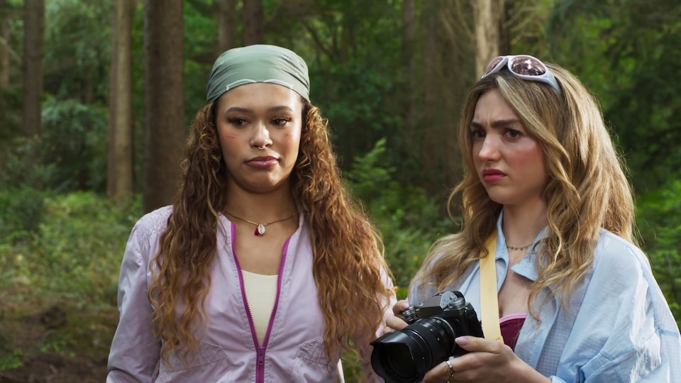 Two women standing in a forest; one holds a DSLR camera, both look puzzled.