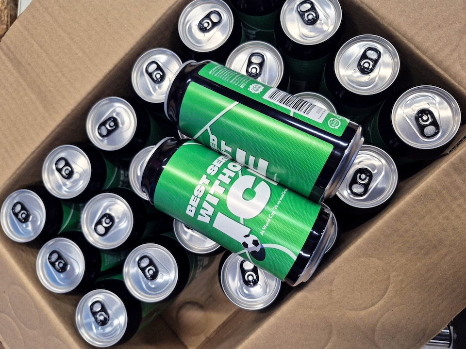 ICE | Muse by Clios Box filled with green-labeled energy drink cans, stacked in a cardboard carton; two cans prominently show a soccer-ball graphic and pull-tabs visible on top surfaces.
