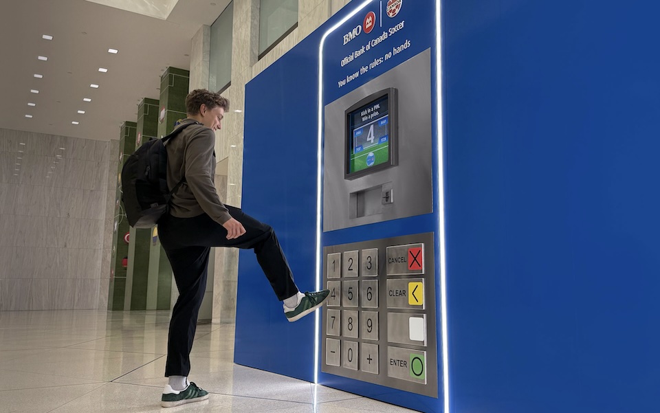 Young man with a backpack kicks a large blue ATM-style kiosk with keypad and screen in a modern lobby.