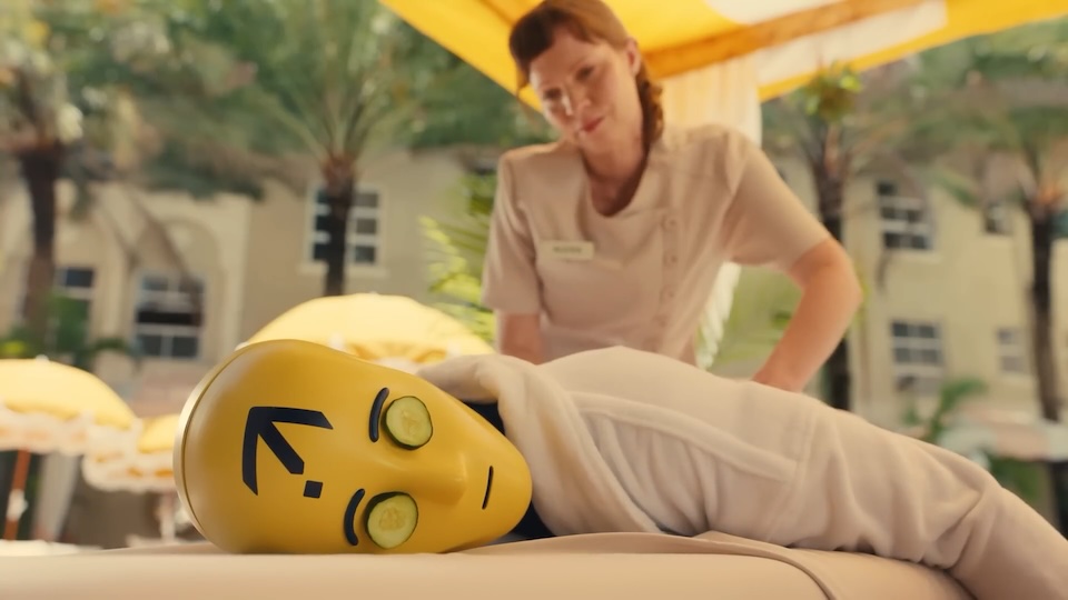 Spa staff adjusts a yellow face prop lying on a massage table with cucumber slices over its eyes for a relaxing treatment.