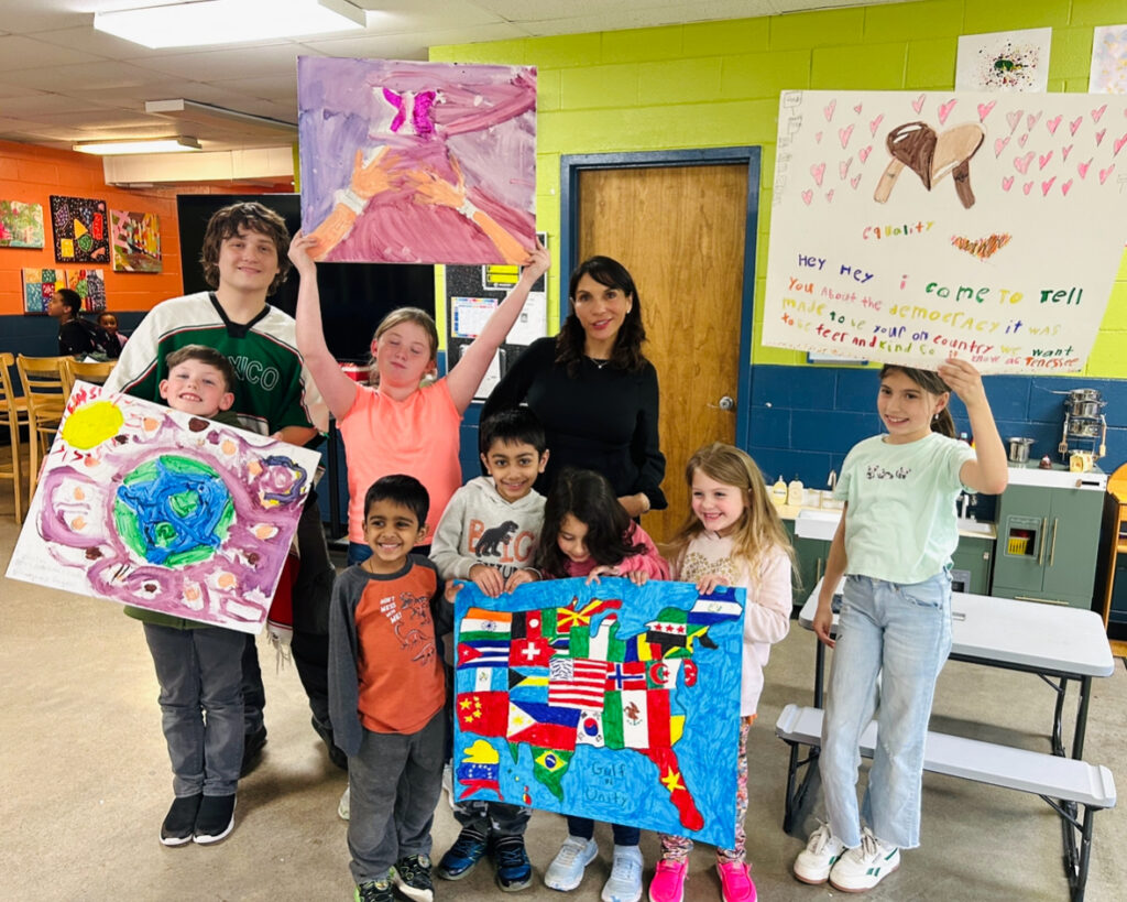 Group of smiling children and a teacher posing with colorful paintings in a bright art room.
