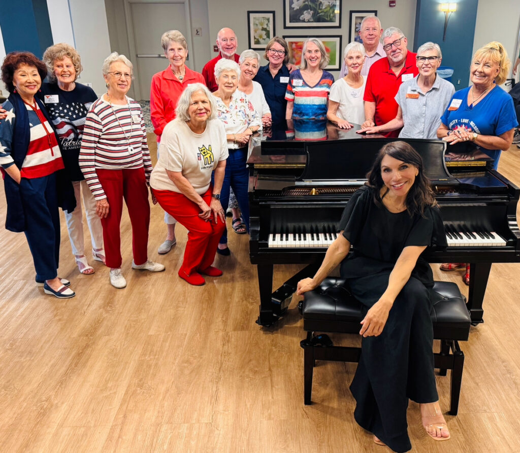 Smiling seniors gather around a black grand piano as a female pianist in a black dress sits at the keyboard.