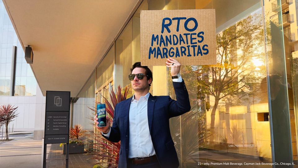 Man in a blue suit and sunglasses raises a cardboard sign reading 'RTO MANDATES MARGARITAS' while holding a Cayman Jack malt beverage outside a glass-front building.