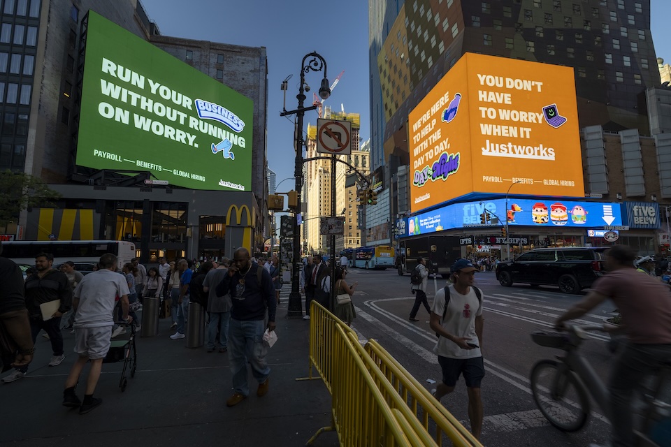 Busy Times Square intersection at dusk with large green and orange billboards and pedestrians crossing the street.