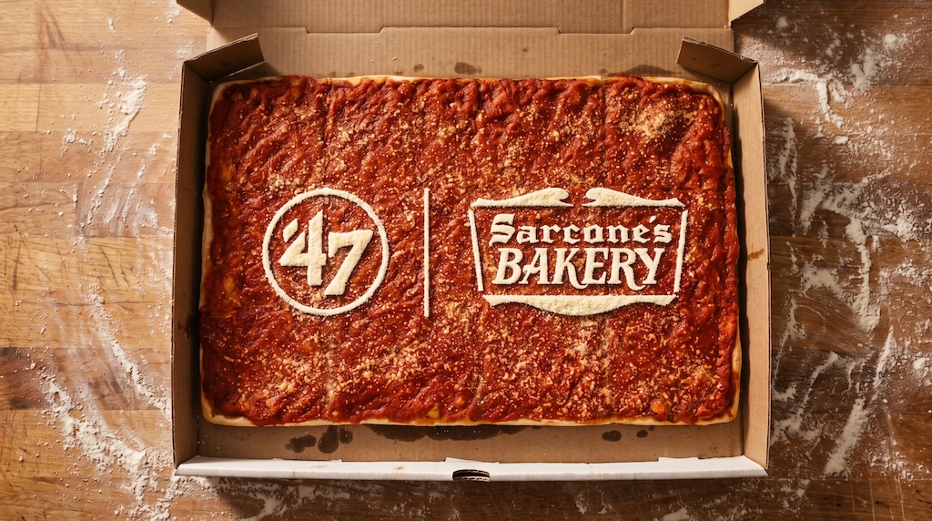 Rectangular pizza in an open box on a flour-dusted wooden table; the sauce surface bears a white baked logo reading '47' and 'Sarcone's Bakery'.