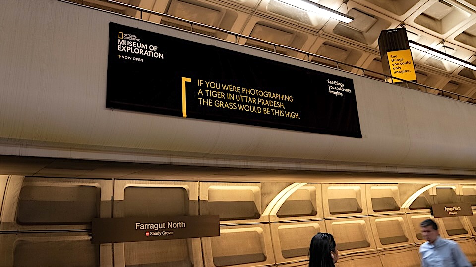 Subway platform with a large National Geographic ad banner about a tiger in Uttar Pradesh and Farragut North station signs visible on the wall.