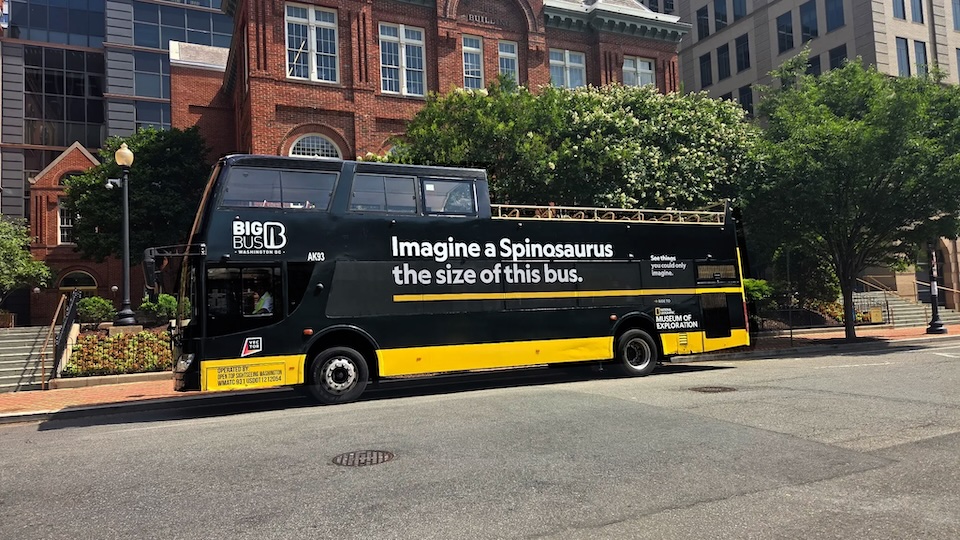 Black double-decker tour bus with a yellow base parked on a city street; advert reads 'Imagine a Spinosaurus the size of this bus.'