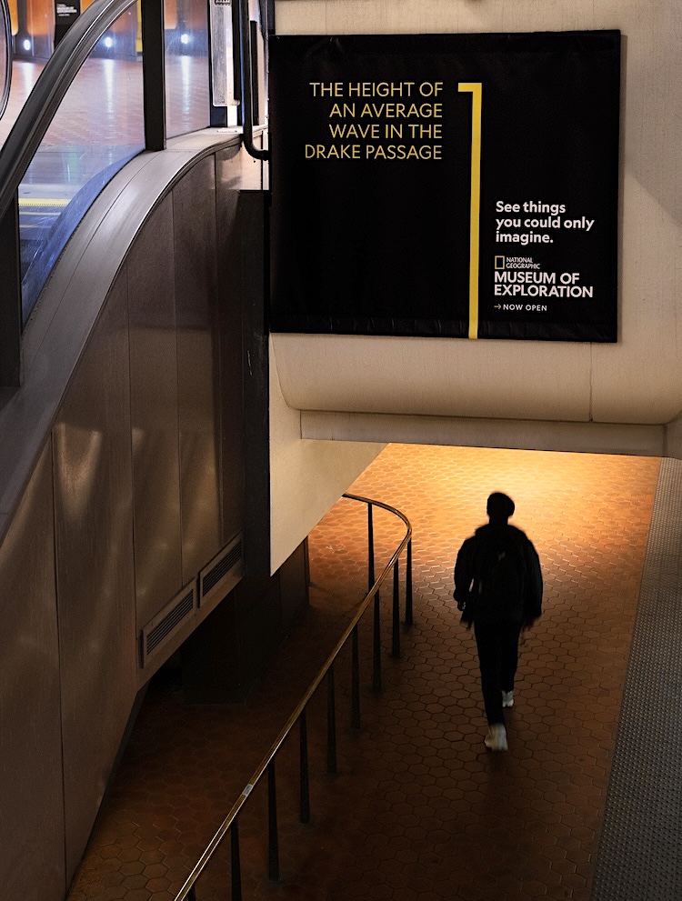 A person walks down orange-tiled stairs in a subway station beside a large black poster advertising the National Geographic Museum of Exploration about Drake Passage waves.