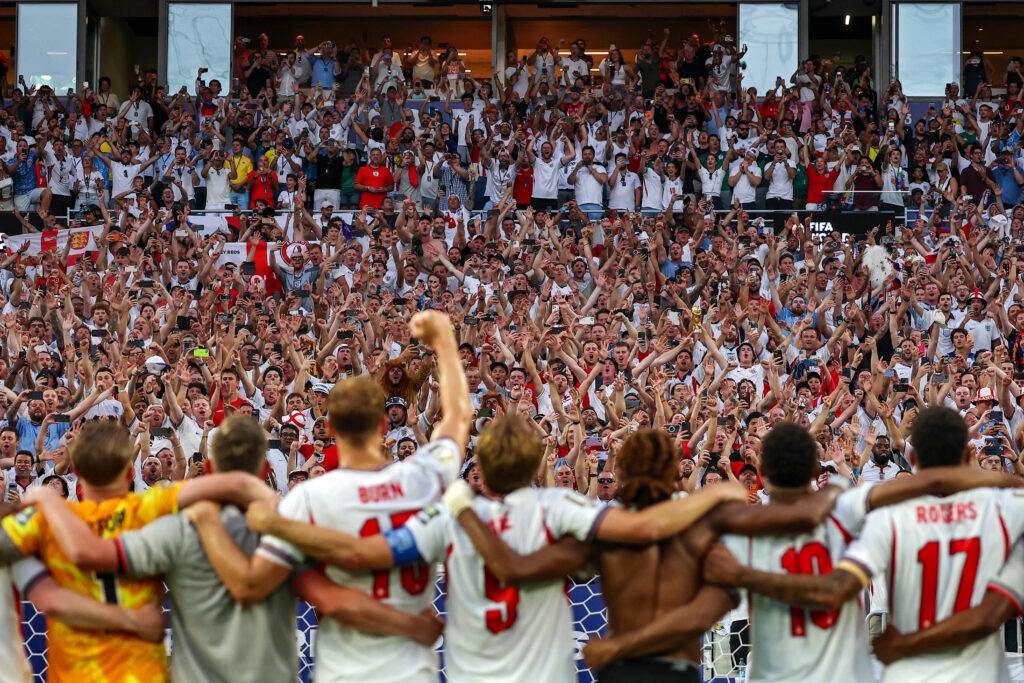 Soccer players hug in a circle as a huge cheering crowd films and chants in white jerseys behind them near a goal.