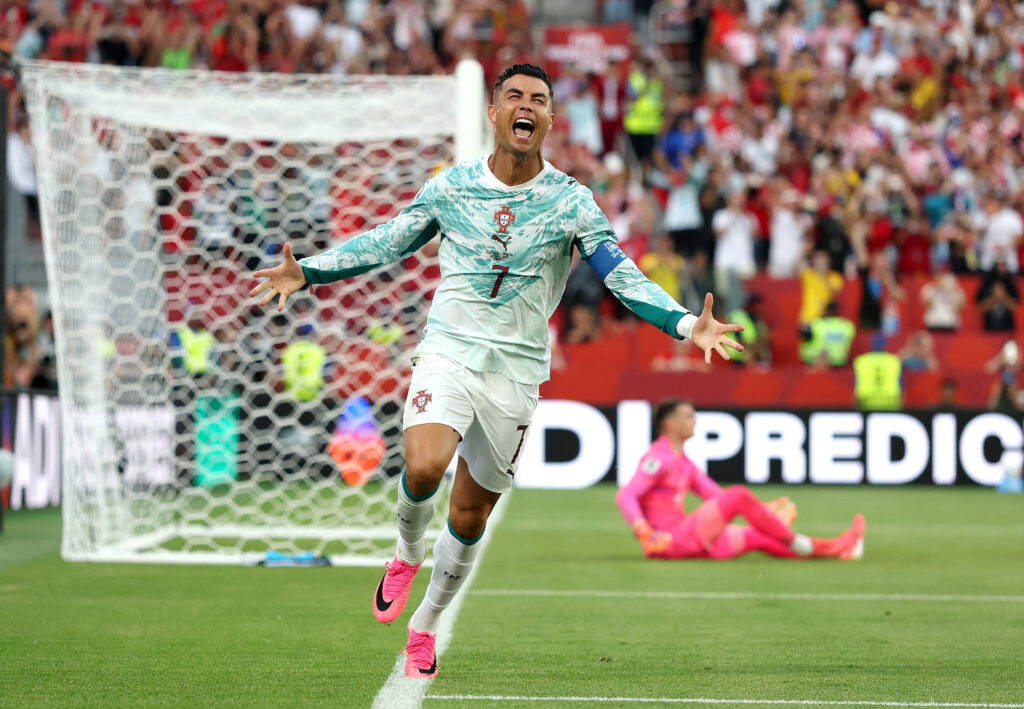 Portugal captain Cristiano Ronaldo celebrates a goal with arms outstretched near the goal, crowded stadium behind him.
