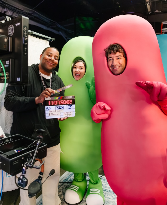 Crew members pose with two costumed mascots (green and pink) on a film set, holding a clapperboard and smiling at the camera.
