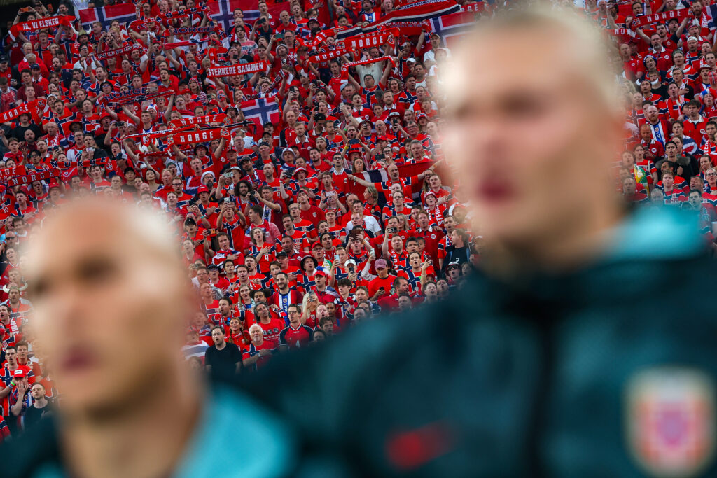 Large crowd of sports fans dressed in red, waving scarves and Norwegian flags at a stadium, with two blurred figures in the foreground