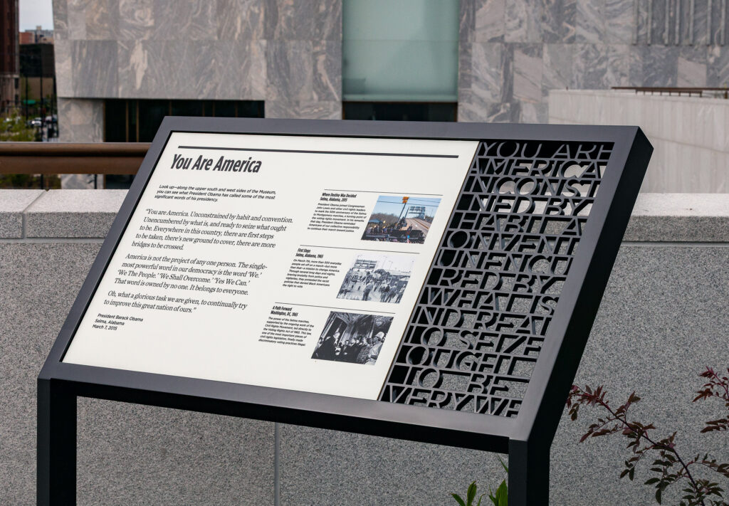 OPC_Selma-panel©Manual | Muse by Clios Informational exhibit panel titled 'You Are America' with text blocks and small images mounted on a dark frame in front of a stone wall.