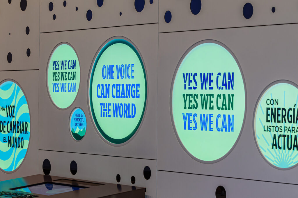 OPC_Exhibit-buttons©Manual | Muse by Clios Circular illuminated panels on a wall displaying the slogans: 'YES WE CAN' and 'ONE VOICE CAN CHANGE THE WORLD' in blue text on green backgrounds, part of an exhibition.