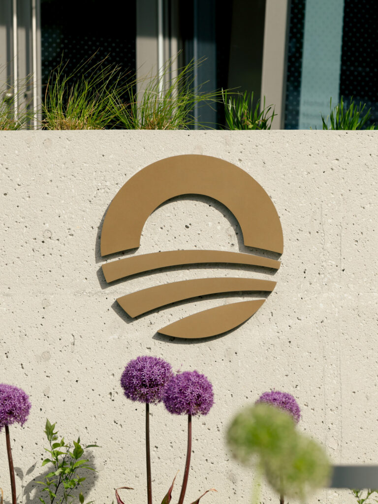 OPC_rising-sun©Manual | Muse by Clios Concrete wall with a gold semicircular metal sculpture, grasses above, and purple allium flowers in the foreground.