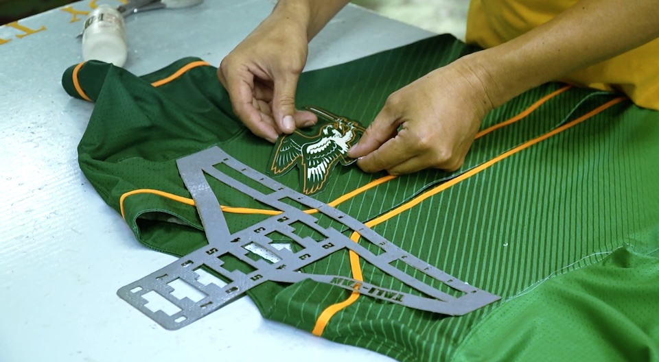 Close-up of hands sewing a patch onto a green athletic jersey on a light blue work surface, with a measuring template nearby.