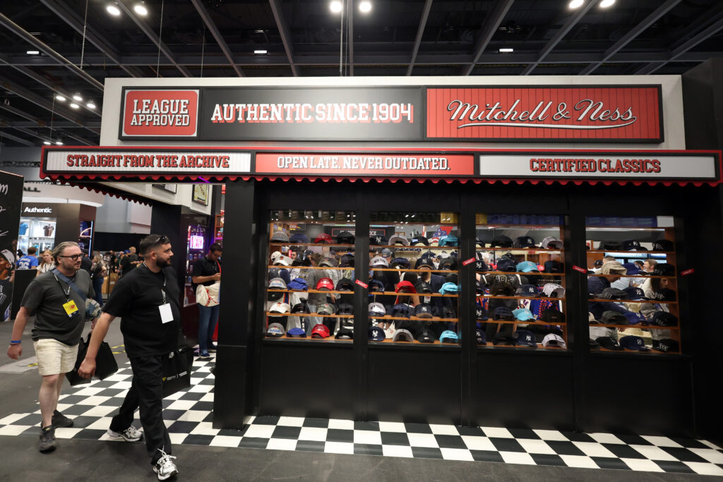 Convention-style hat shop booth with shelves of baseball caps and bold'Mitchell & Ness' signage overhead, checkered floor visible.