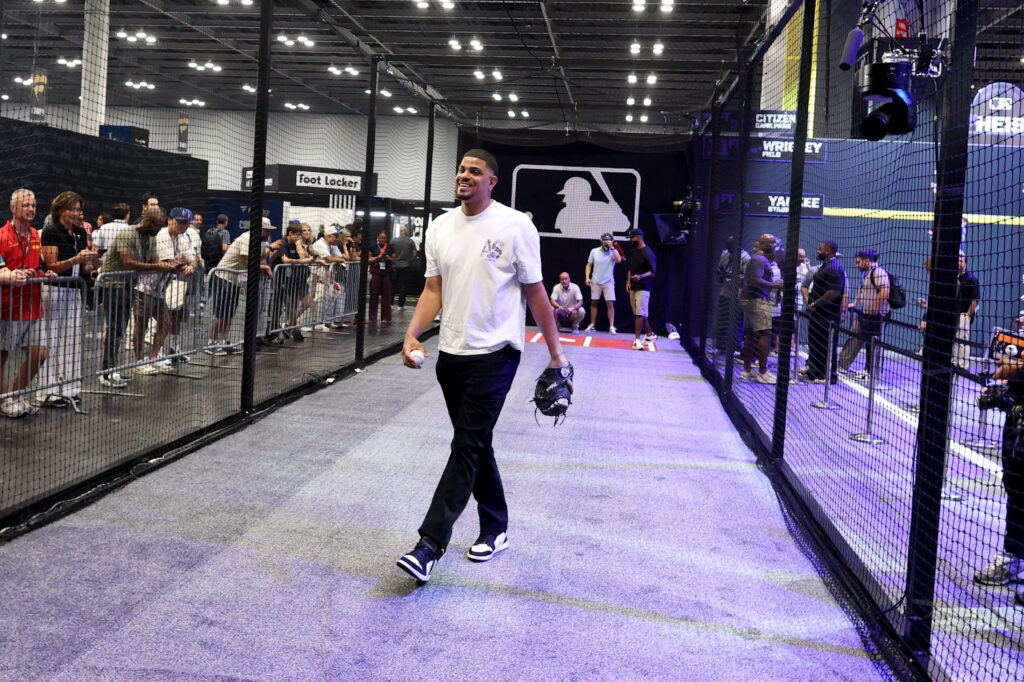 Man in a white shirt walks through an indoor batting cage while spectators watch from behind nets.