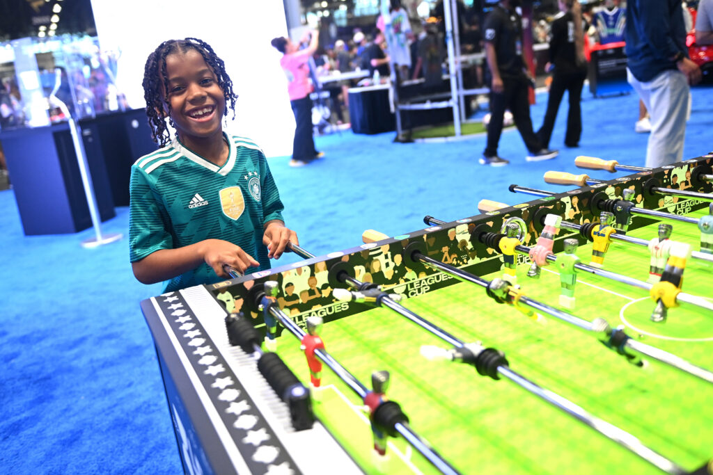 Smiling girl in a teal soccer jersey plays foosball at an indoor event.