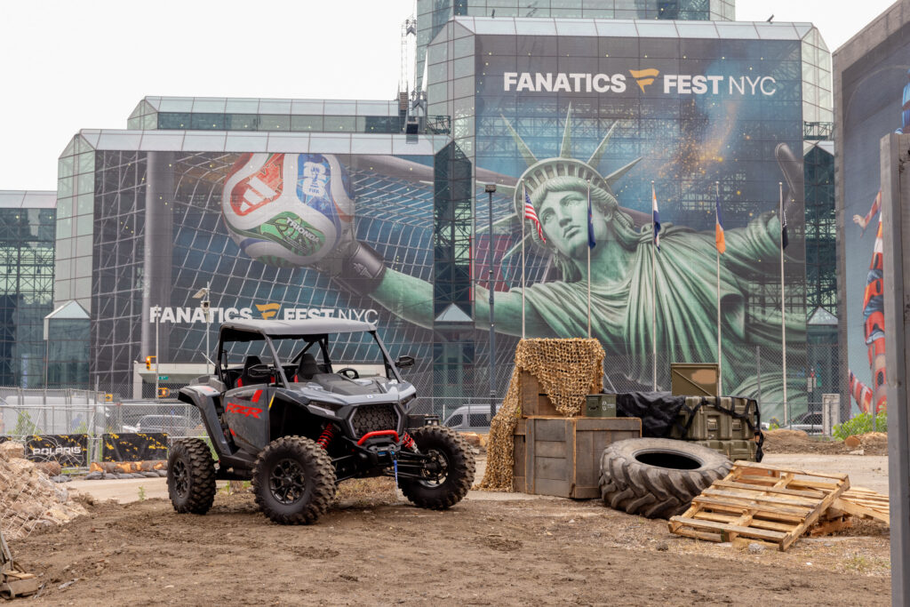 Construction site in front of a glass building with a Fanatics Fest NYC banner; a Polaris RZR off-road vehicle sits in the foreground.
