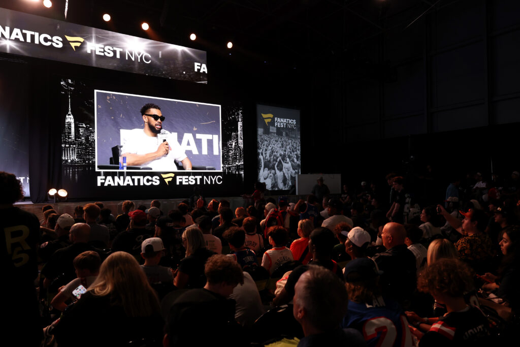 Speaker on stage at Fanatics Fest NYC, microphone in hand, with a large screen displaying him and a cheering crowd in front.